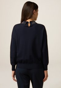 Suéter azul marino con escote redondo, presenta un cuello con cuentas y un detalle de apertura en la parte posterior. Textura suave y corte relajado, los puños son acanalados.