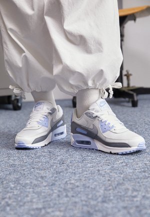 Persoon die witte joggerbroek en witte Nike Air Max sneakers met lichtblauwe en grijze accenten draagt, staand op een met tapijt beklede vloer.