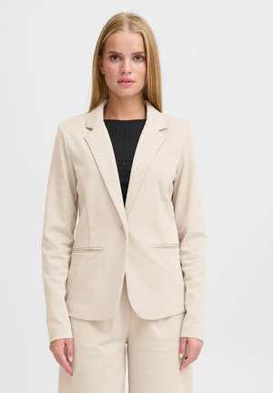 Lyshåret kvinde iført beige skræddersyet blazer og matchende bukser over en sort ribbet top, stående foran en ensartet hvid baggrund.