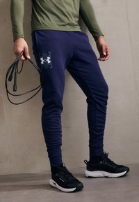 Jūras zili sporta bikses ar pieguļošu dizainu, kurām kreisajā augšstilbā ir Under Armour logo; apvienotas ar melniem sporta apaviem un lecamaukla.