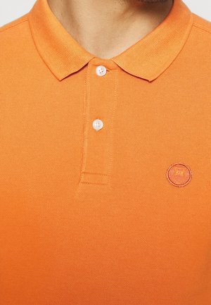 Mężczyzna w pomarańczowej koszulce polo z dwoma białymi guzikami i naszywanym logo sowy na lewej piersi.