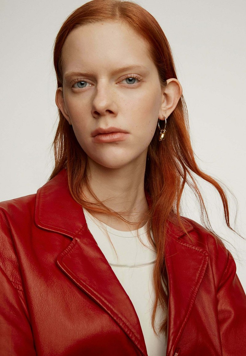 Chaqueta de cuero roja con solapas, superpuesta a una blusa blanca. Cabello largo y liso de color rojo, y un pequeño pendiente de aro con un charm.