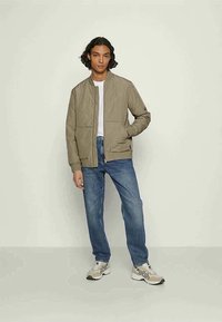 Beige quilted bomber jacket with a round collar, front zipper, and two side pockets, paired with light blue jeans and neutral sneakers.