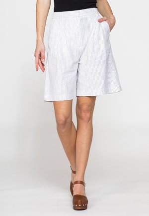 Donna che indossa pantaloncini bianchi a righe sottili fino al ginocchio e zoccoli marroni con tacco, in piedi con una mano in tasca su sfondo semplice.