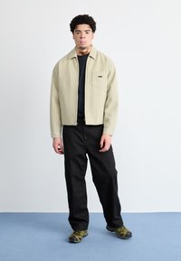 Jachetă crem deschis, cu dungi, cu fermoar și un buzunar pe piept și logo-ul "OBEY", purtată cu pantaloni negri și adidași verzi/negru.