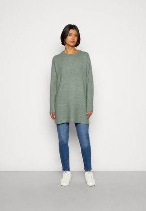 Pull en tricot à manches longues, de couleur vert clair, avec un col rond, porté sur un jean skinny bleu et des baskets blanches, offrant une coupe décontractée.