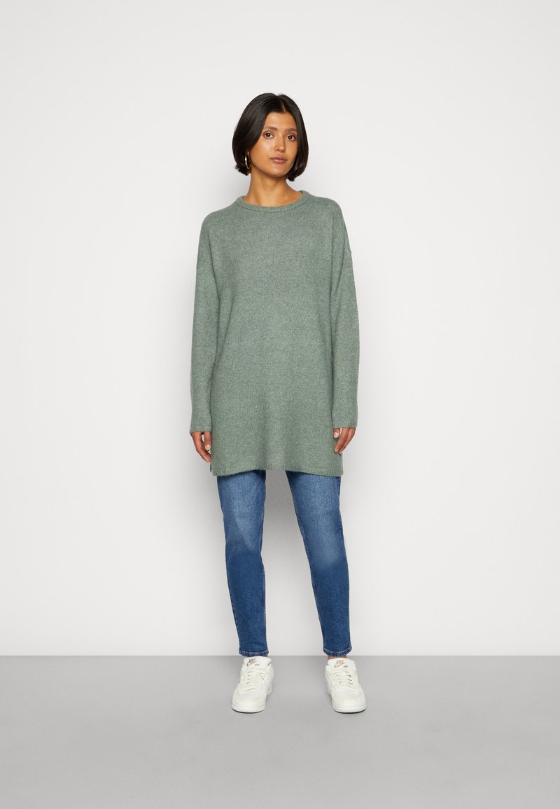 Sweater de malha de mangas compridas, verde claro, com decote redondo, usado sobre jeans justos azuis e ténis brancos, apresentando um ajuste relaxado.