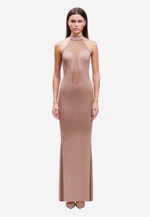 Vrouw met steil bruin haar draagt een nauwsluitende mouwloze taupekleurige jurk met meshpanelen en een hoge halslijn, staand tegen een witte achtergrond.