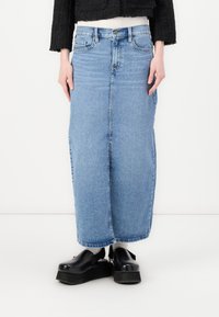 Jupe maxi en denim bleu clair avec une silhouette droite, taille haute et fente à l'avant. Assortie à des chaussures à plateforme noires.
