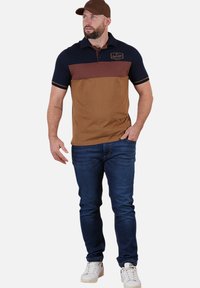 T-shirt polo avec col bleu marine et manches courtes, présentant une rayure horizontale marron et un corps beige. Porté avec un jean foncé et des baskets blanches.