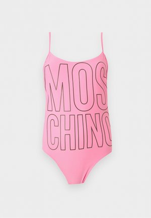 Młoda kobieta stojąca boso, ubrana w różowy jednoczęściowy strój kąpielowy z dużym napisem "MOSCHINO".