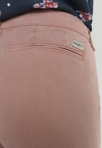 Růžové kalhoty vyrobené z textilního materiálu, s zadní kapsou a malou značkovou visačkou na pase. Design je prostý a klasický.