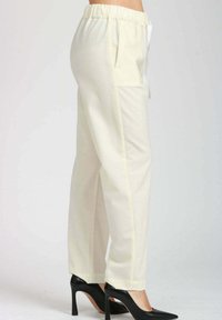 Pantaloni a vita alta color crema con fascia elastica in vita e tasche laterali, indossati con tacchi a stiletto neri a punta su uno sfondo chiaro.