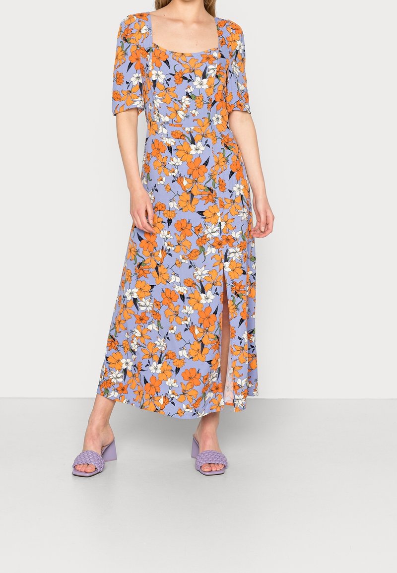 Femme portant une robe lilas avec un imprimé floral orange et blanc, encolure carrée, manches mi-longues, fente latérale et sandales à talons lilas.