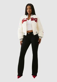 White faux fur bomber jacket with red embroidery, black and white striped cuffs, paired with black flared trousers and red pointed heels.