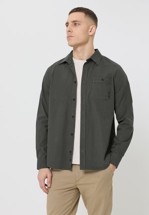 Mann trägt ein dunkelgraues Hemd über einem weißen T-Shirt und beigen Hosen, posiert mit einer Hand in der Tasche und schaut nach links, vor einem schlichten Hintergrund.