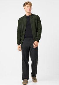 Młody mężczyzna w ciemnozielonym swetrze zapinanym na zamek, czarnej koszuli, czarnych spodniach i brązowych sneakersach stojący na białym tle.