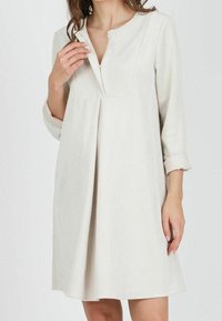 Nainen, joka on pukeutunut polvipituiseen, pitkähiharaiseen kermaisenväriseen corduroy-mekkoon, jossa on piilotettu etuvetoketju ja pussit edessä vyötäröllä.