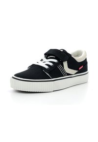 Zapatilla de lienzo negra con cordones blancos y acentos en crema en contraste. Cuenta con una correa de Velcro y una suela de goma texturizada con ranuras horizontales.