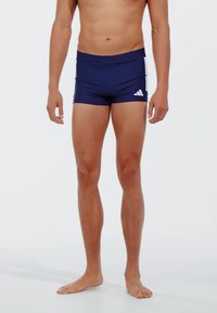Mornarsko plave kupaće gaće s pripijenim dizajnom, izrađene od elastičnog materijala; sadrže bijeli Adidas logo na lijevom kuku.