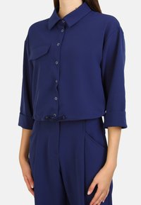 Mörkblå kortärmad skjorta med knappar, bröstficka, uppvikta ärmar och justerbar dragsko i midjan. Tillverkad av mjukt tyg.