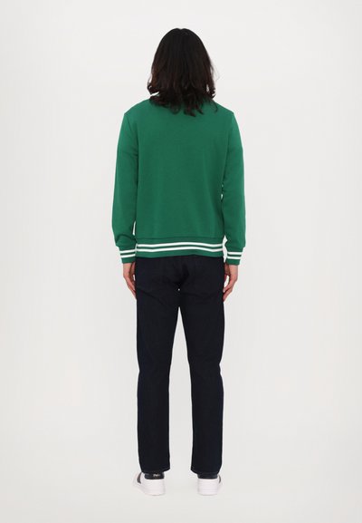 Grønn hettegenser med hvite striper ved mansjettene og kanten, kombinert med mørkeblå jeans og hvite joggesko, sett fra baksiden.