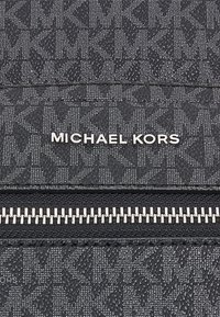 Portefeuille en tissu texturé noir avec un motif de logo répété, marquage "Michael Kors" en argent, et une fermeture éclair avec des dents en métal.