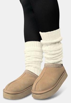Hellbraune Wildleder-Slipper mit einer strukturierten geflochtenen Detail am Knöchel, kombiniert mit gemütlichen weißen, gerippten Stulpen über schwarzen Leggings.