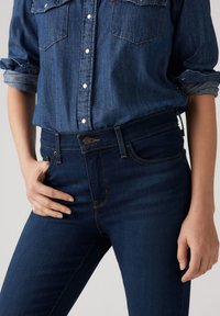 Tummansiniset farkut korkea vyötärö, viisi taskua ja edessä nappikiinnitys. Yhdistetty vaaleansinsiseen denimipaitaan, joissa on käännetyt hihat.