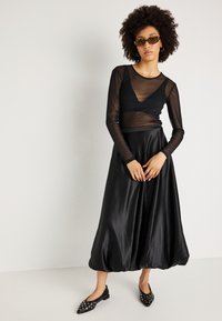 Fekete átlátszó, hosszú ujjú blúz fekete selyem szoknyára rétegződve, amely ballon formájúra bővül. Lapos fekete cipővel viselve, fémes díszítéssel.