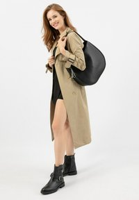 Trench coat beige chiaro con bottoni, borsa a mano in pelle nera e stivaletti neri con fibbie. Il modello si trova su uno sfondo bianco.