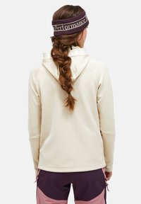 Light beige hooded sweatshirt with a smooth texture. Braided hair and a dark purple headband. Tight-fit cuffs and a subtle branding detail on the back.