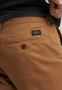 Pantalones de algodón marrón con un bolsillo trasero con un botón marrón y una etiqueta con texto negro, acompañados de una camisa negra lisa.