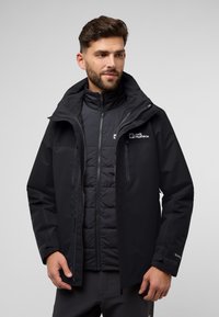 Veste noire imperméable avec capuche détachable, dotée de zips, poches et isolation en baffles en dessous pour chaleur et confort.