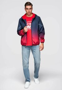 Mladý muž ve zvedavé bundě od námořnické modré po červenou, červeném tričku s potiskem, světle modrých džínách a bílých teniskách stojící na jednobarevném pozadí.