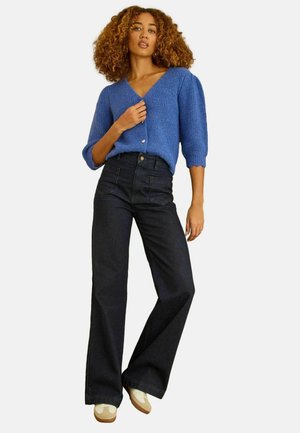 Kiharate juustega naine, kes kannab sinist nööpidega kardigani, kõrge vöökohaga laia säärega musti teksapükse ja beeže tossusid, seisab käsi rinna lähedal.