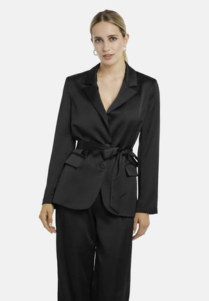 Frau mit blondem Haar, die einen schwarzen Satin-Blazer mit gebundenem Taillengürtel und passende weite Hosen vor weißem Hintergrund trägt.