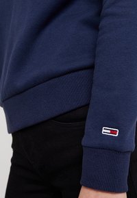 Närbild av en person som bär en marinblå sweatshirt med en liten rektangulär logotyp i röd, vit och svart på ärmsluten.