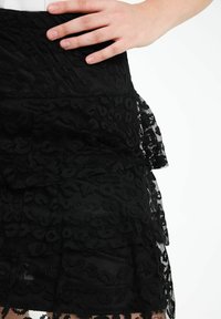 Jupe en dentelle noire avec volants superposés, présentant un motif floral texturé et une taille lisse. Les détails soulignent le design complexe du tissu.