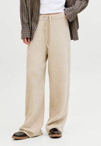 Pantaloni in maglia larghi color beige con vita regolabile indossati con scarpe nere, una maglietta bianca e una camicia a quadri aperta.