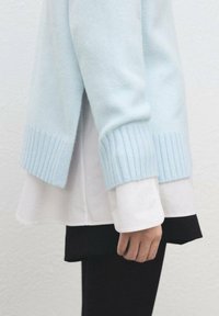 Svetlo modra pletena majica z rebrastimi manšetami, nošena čez belo srajco in črni spodnji del. Ima razporek na robu in mehko teksturo.