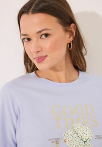 Lichtpaarse langemouwen t-shirt met gele tekst "GOEDE TIJDEN" en een illustratie met bloemmotieven. Zachte stofstructuur, relaxte pasvorm.