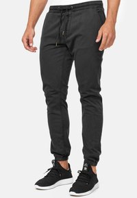 Homme portant un pantalon de jogging noir avec des poignets élastiques et une taille à cordon, associé à des baskets Adidas noires et blanches.