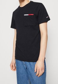 T-shirt en coton noir avec une poche sur la poitrine ornée de rayures rouge, blanc et bleu ; encolure ronde et manches courtes.