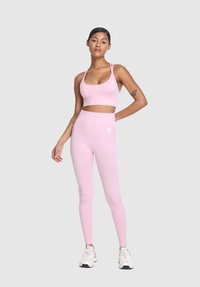 Ženska v roza športni modrček in leginsih, z belimi supergami, stoji s roko na bok proti svetlo sivi podlagi.