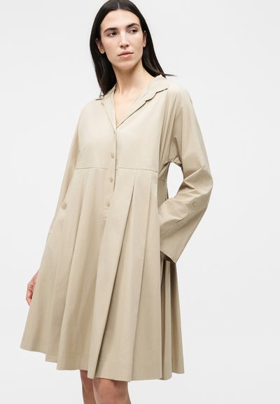 Γυναίκα με μακριά σκουρόχρωμα μαλλιά, φορώντας ένα φαρδύ, μπεζ φόρεμα με μακριά μανίκια, κουμπιά στο μπροστινό μέρος και πλισέ φούστα, στέκεται μπροστά σε απλό φωτεινό φόντο.
