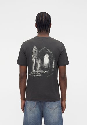 Mann mit Cornrow-Zöpfen, der ein schwarzes T-Shirt mit gotischem Kathedralaufdruck und dem Text "Es gibt ein Licht, das niemals verschwindet" auf dem Rücken trägt, kombiniert mit blauen Jeans.