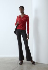 Red draped top with long sleeves, paired with flared black jeans. Model wears black platform sandals. Minimalist background.