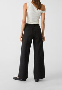 Zwarte wijde spijkerbroek met hoge taille, voorzien van twee achterzakken en een rafelrand. G gedragen met een witte schoudertop en zwarte platte schoenen.