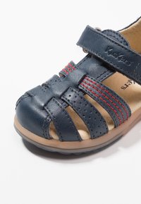Sandal de cuero azul marino con diseño de punta abierta, tiras entrecruzadas, acentos de costura roja y una suela de goma beige flexible.